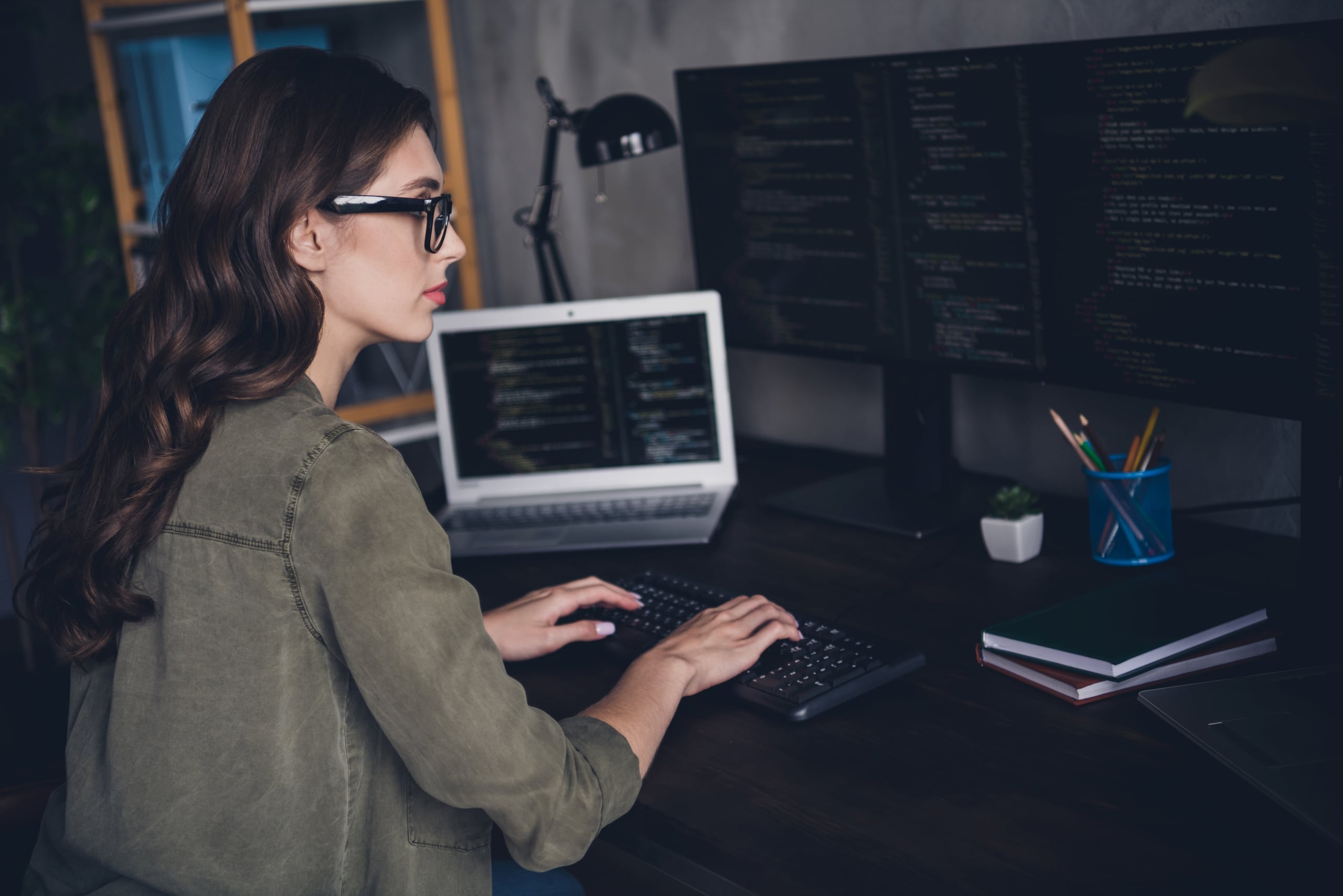 Photo of confident lady hacker wear eyeglasses writing binary code at modern desk indoors with multiple workplace workstations