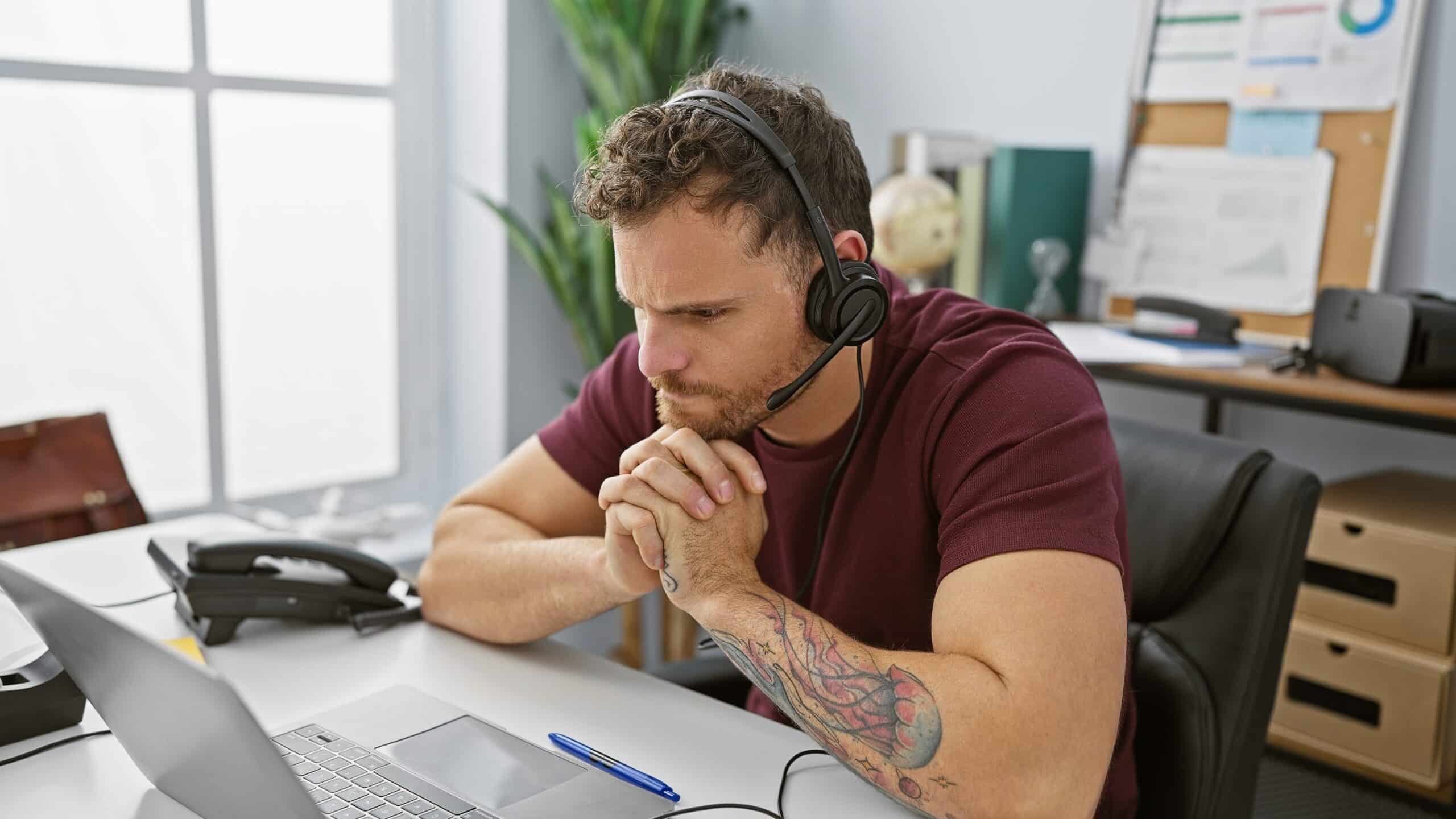 A focused dark-haired man with a beard and tattoo works indoors at an office wearing a headset in front of a laptop.